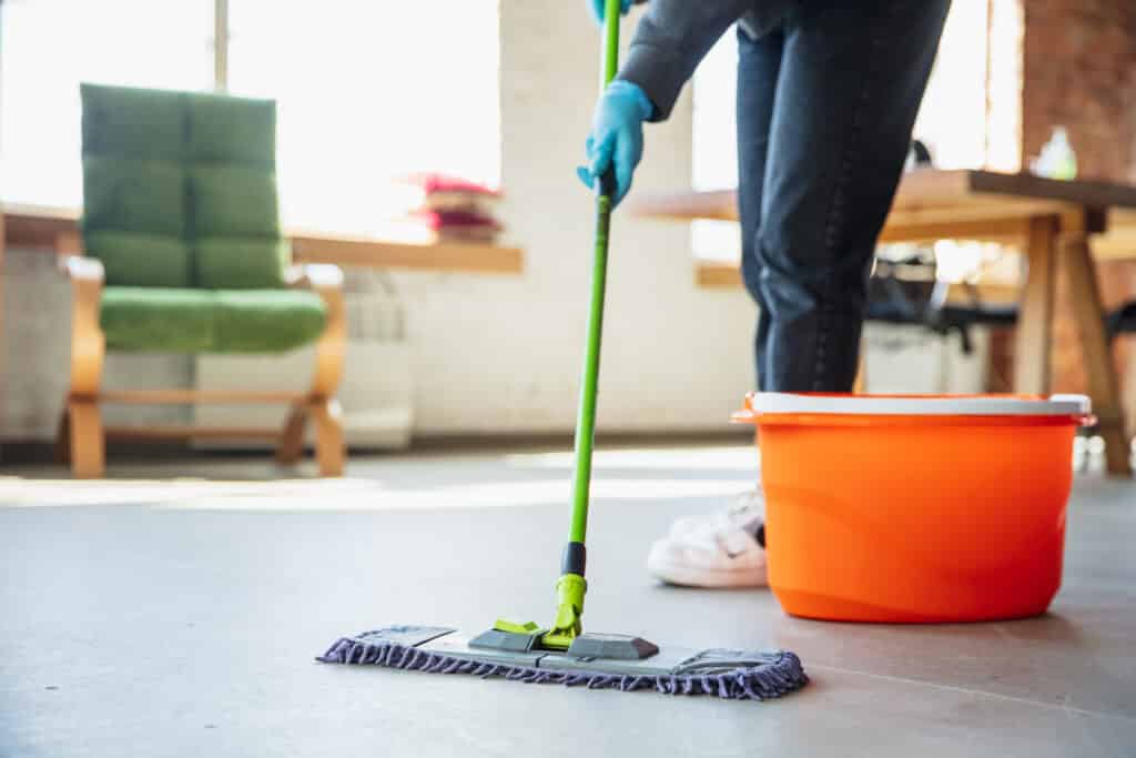 The image shows an SCS Group Commercial Cleaner mopping the floor diligently in a room. They use a mop and bucket, displaying dedication and precision. A green chair adds color to the space, while white sneakers and an orange juice bottle suggest a temporary break. The cleaner's meticulous effort ensures a fresh and pristine environment.