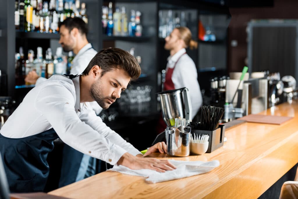 A diligent bartender in a red apron cleans the bar counter. A sleek silver blender and freshly brewed coffee machine add to the ambiance. A barber tends to a gentleman's haircut. A white bowl holds a cup of steamy coffee, while a green toothbrush adds a colorful touch.