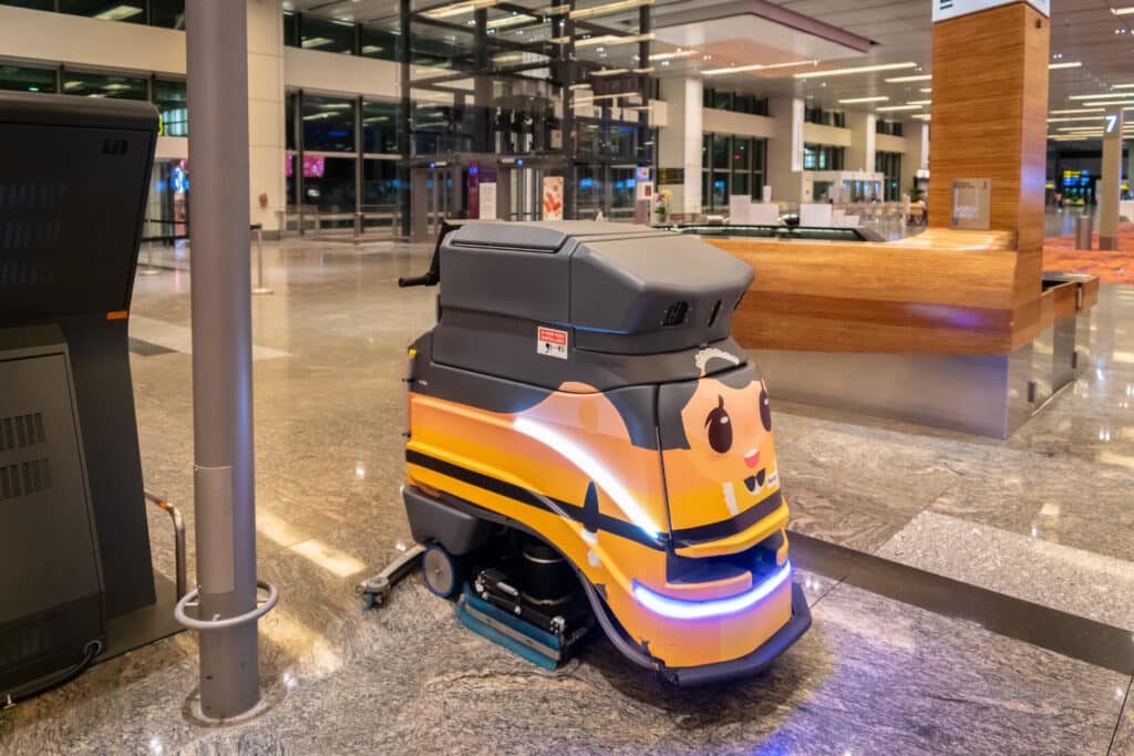 A sleek cleaning robot operates in a bustling airport lobby. A futuristic interface screen displays interaction options. A man observes outside. The modern ambiance features a black TV and bustling travelers.