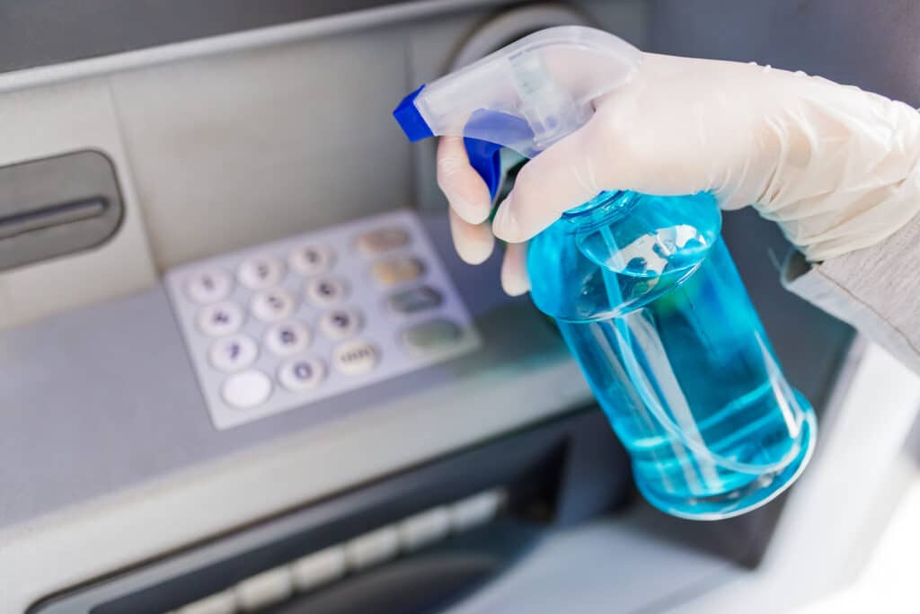 A Bank cleaning personnel in blue gloves sprays a mist of blue liquid onto an ATM machine, creating a sense of mystery and urgency in the air.