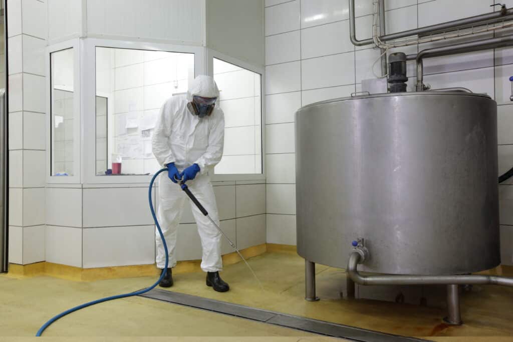 A industrial cleaner in a white protective suit cleans a stainless chemical steel tank with a blue hose. He mops the floor, wearing a mask for protection. A skateboarder glides through a subway station, pipes adorn a wall, combining cleanliness and urban vibrancy.