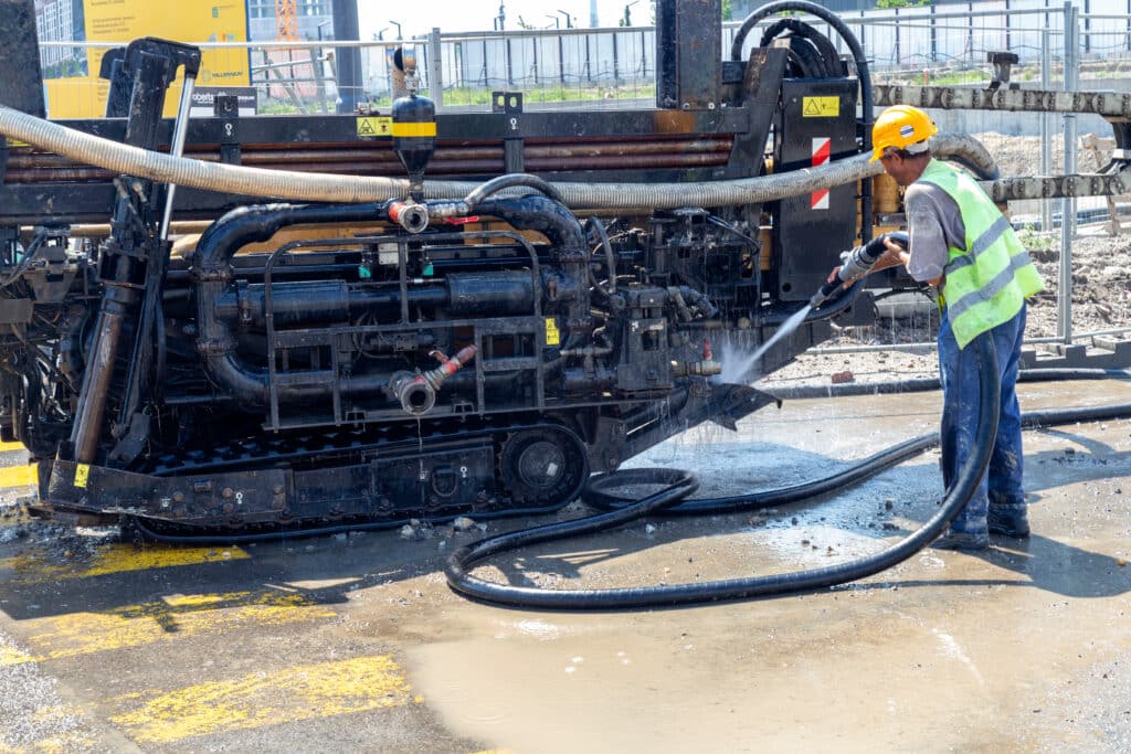 An industrial cleaner in a yellow vest and hard hat uses a hose to clean a machine on a mining site.