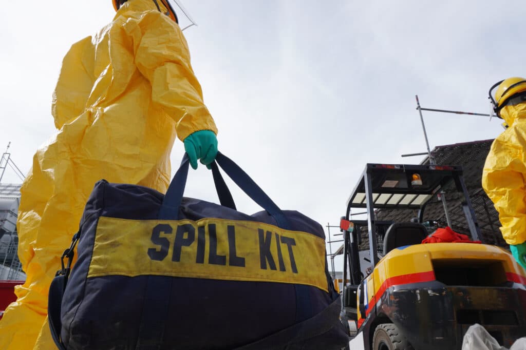 Two people in yellow suits carry a "Spill Kit" bag, while a forklift, SPC-branded bags, and a person in a yellow helmet complete the scene of safety equipment.