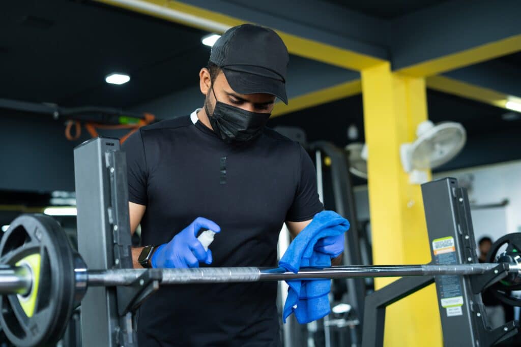 A determined commercial cleaner in a gym wears a mask and gloves while lifting a barbell, surrounded by exercise equipment. His blue towel and spray bottle underscore his commitment to hygiene and safety.