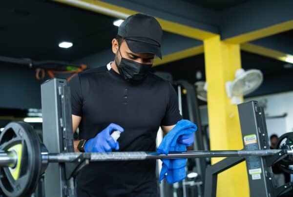 A focused gym cleaner wears a mask and gloves. They lift a barbell and are surrounded by exercise equipment. His blue towel and spray bottle underscore his commitment to hygiene and safety.