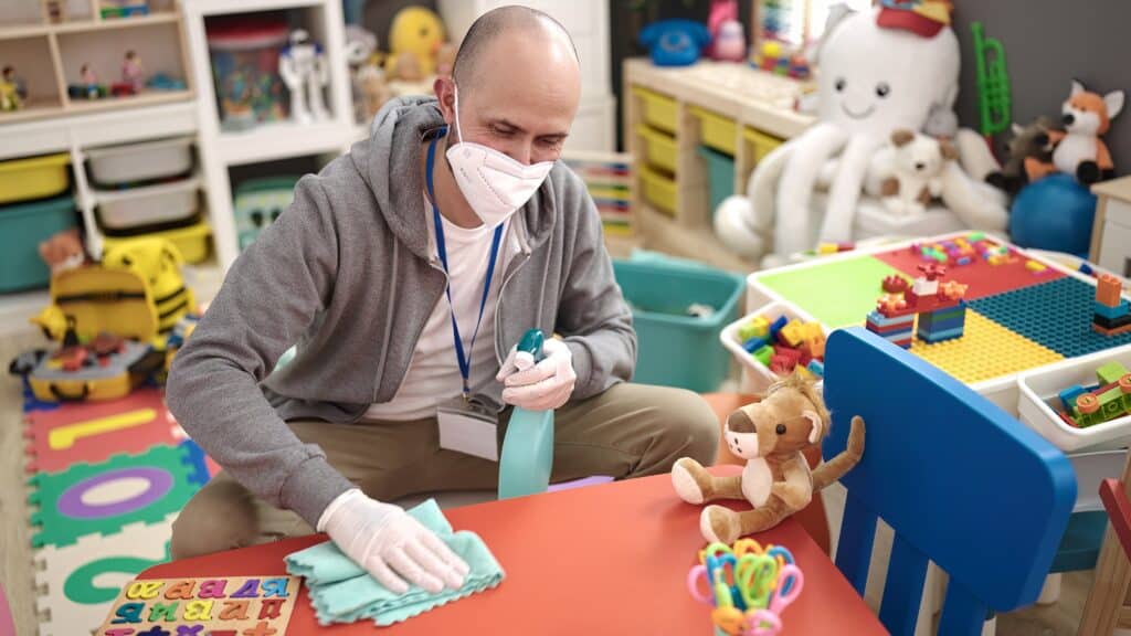 A masked commercial cleaner meticulously cleans toys in a childcare room. Teddy bear toys, an alphabet puzzle, and a lion and octopus plushies adorn the scene.