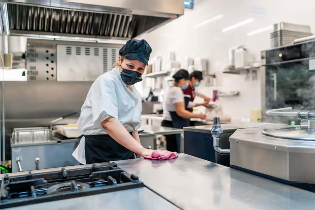 Two women in masks work in a bustling restaurant kitchen. Modern appliances, a gas stove, and skilled staff create an efficient and vibrant atmosphere.