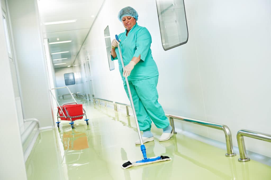 A commercial hospital cleaner in scrubs cleans the hospital hallway with a mop. A red bucket rests nearby. Doctors, nurses, and patients move purposefully. A colorful window picture offers respite. Collaboration is evident, while a gleaming sink handle underscores sanitation.