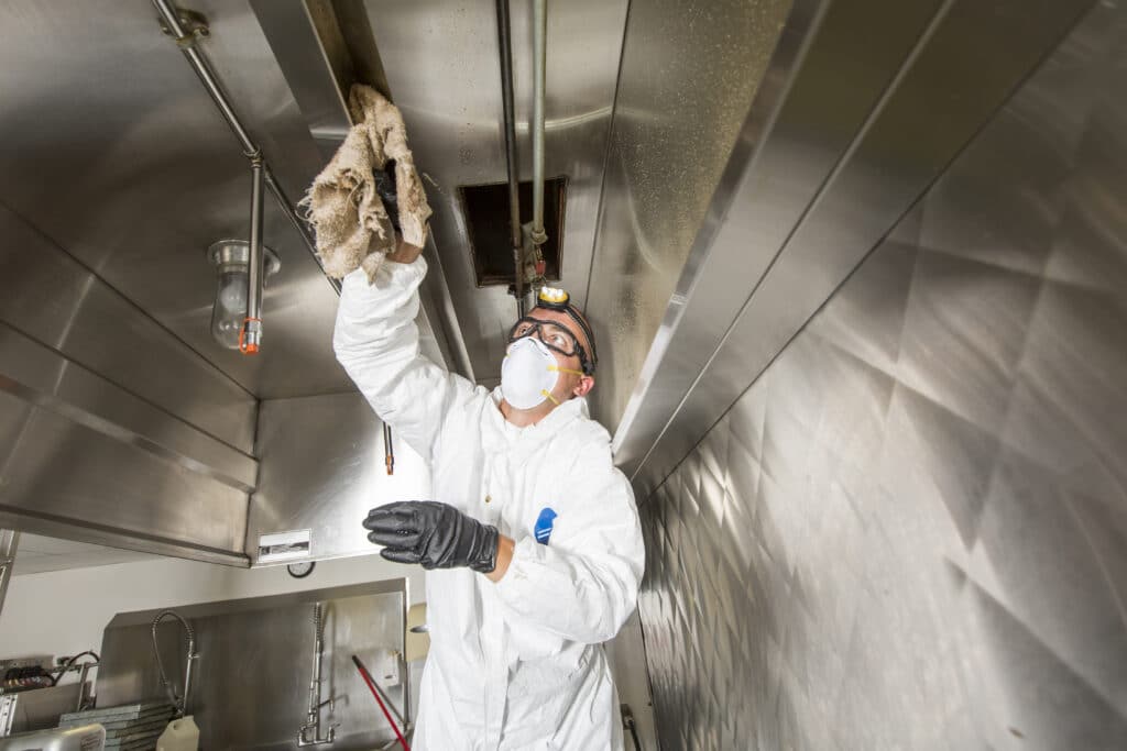 A young man in a white protective suit works on the ceiling of an industrial kitchen, holding a plush stuffed animal. Stainless steel surfaces, safety gear, and a light fixture create an atmosphere of focus and comfort.