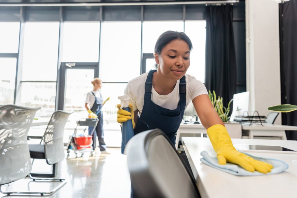 Smiling,Bi-racial,Woman,Cleaning,Office,Desk,With,Rag,Near,Colleague